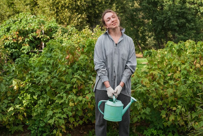 A woman holds a watering can while wearing gardening gloves, smiling in her garden