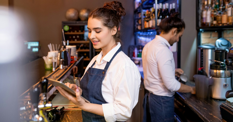 A waitress stands behind the bar looking at a tablet, while a colleague works behind her