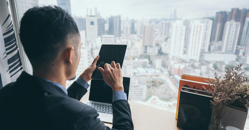 A businessman, seen from behind, works on his tablet with his laptop and a view of the city skyline in front of him