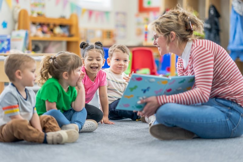 A preschool teacher sits on the floor of her classroom with a small group of students as she reads them a book