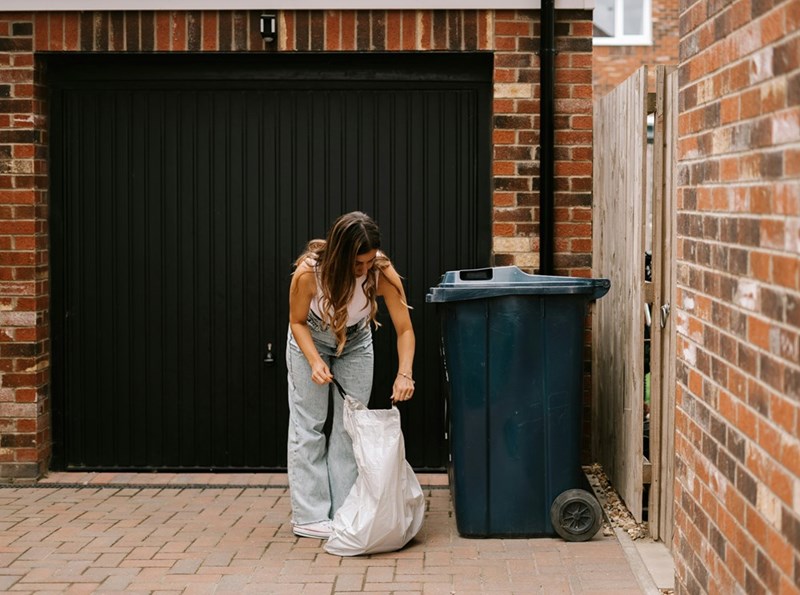 A woman collecting her trash and throwing it in a neighbor's trash bin.