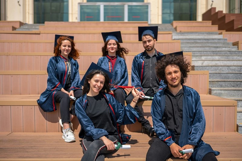 High school students smile on the steps of their school wearing cap and gown.