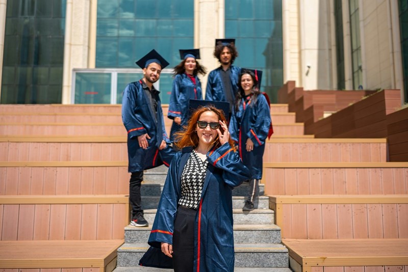 A high school senior poses in sunglasses, cap, and gown surrounded by four friends.