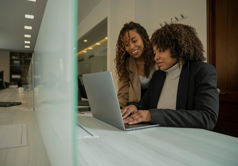 One female employee assists another as they work in front of a glass partition in the office.