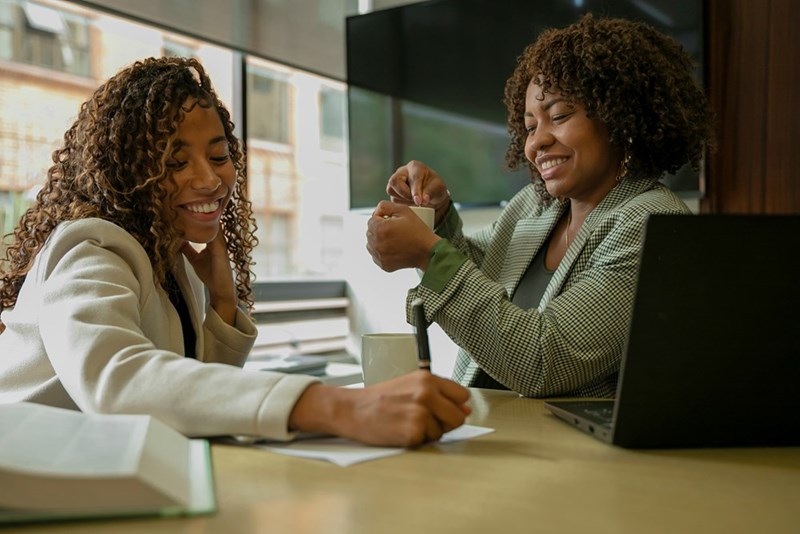 Two female coworkers collaborate in a conference room while exchanging polite smiles.