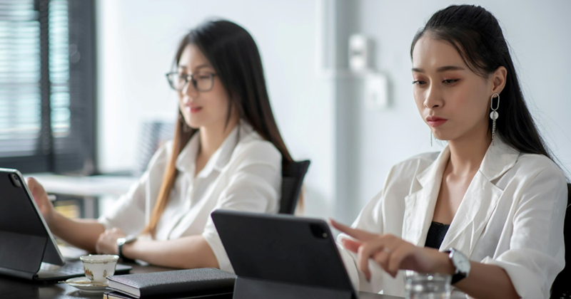 Two female employees sit together working on tablets, with one pointing at her tablet's screen