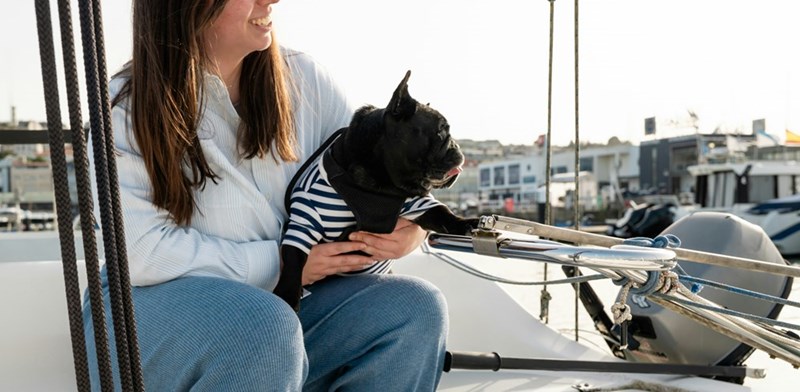 Woman sitting on the pope of a boat with a black bulldog wearing a striped black and white outfit.