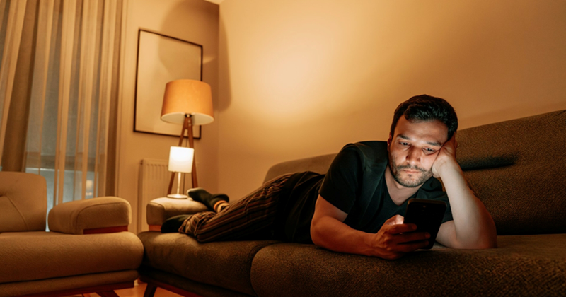 A man lies on a couch, looking at his phone in the evening