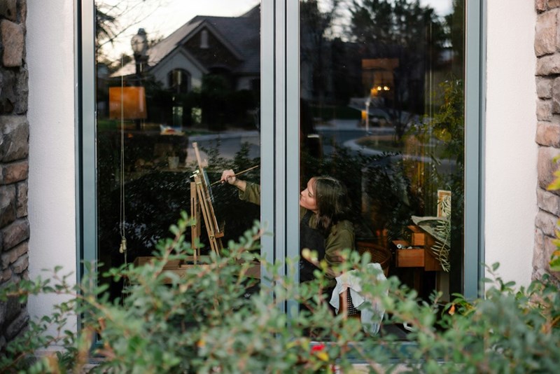 A woman painting a canvas seen from the outside of a house through a window surrounded by plants.