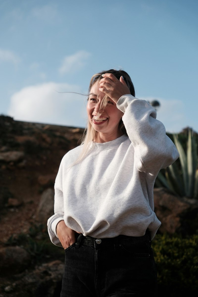 Woman smiling outdoors in a white sweatshirt with wind-blown hair under a bright blue sky.
