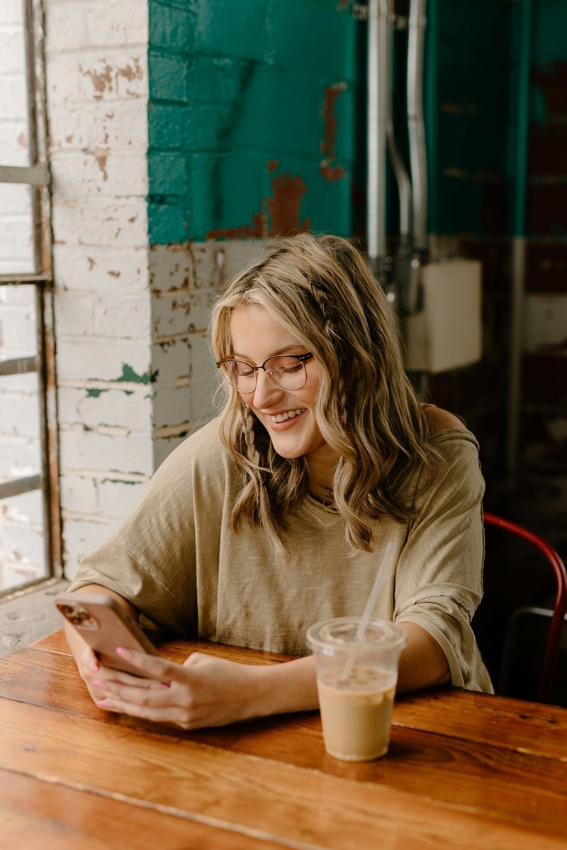 Woman smiling at her phone in a cafe, wearing glasses and sitting beside an iced coffee.