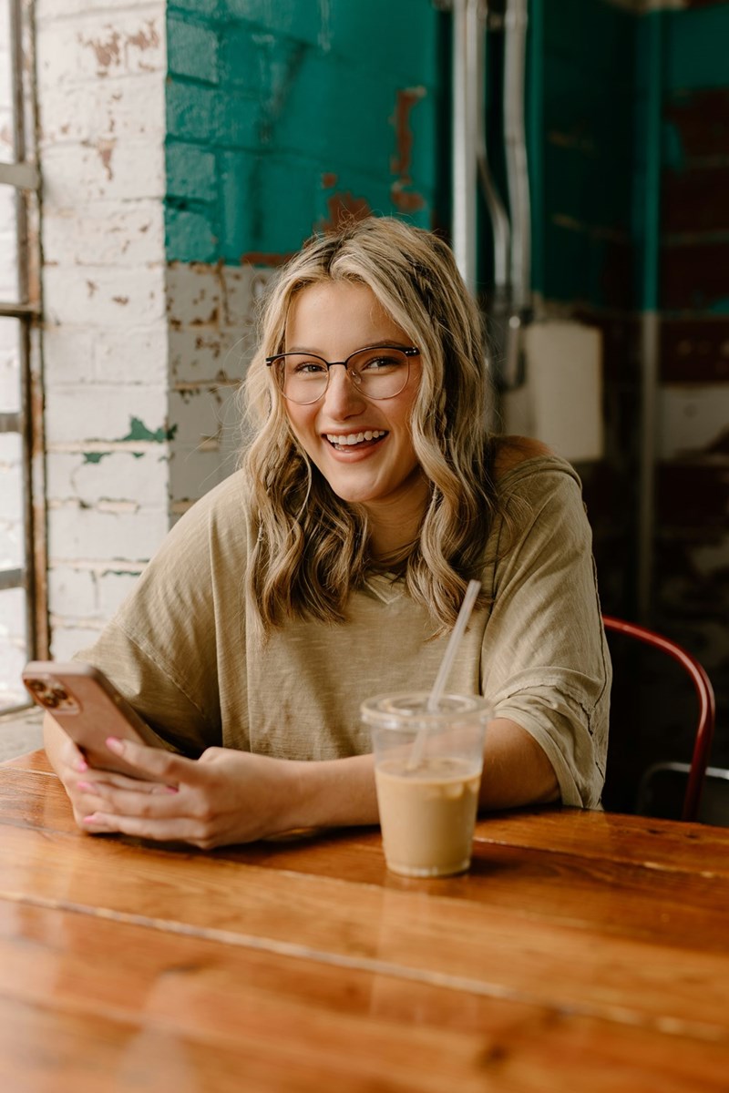 Smiling woman with glasses using her phone at a cafe table beside an iced coffee drink.