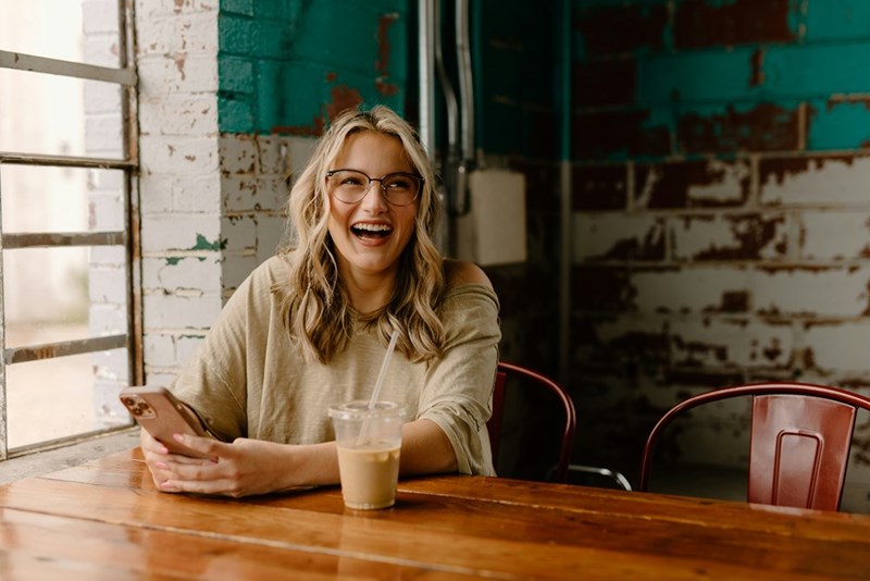 Woman laughing in a cafe while holding her phone, seated beside an iced coffee on a wooden table.