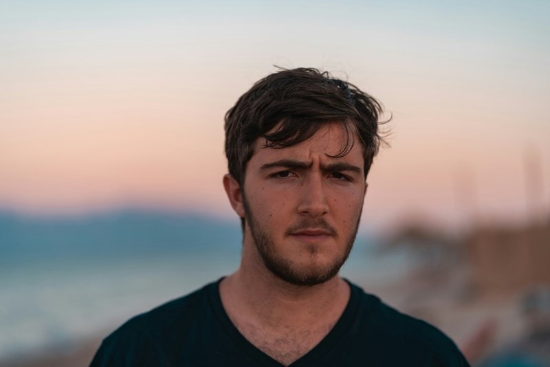 Young man with a beard standing by the water at sunset, wearing a dark shirt and looking serious.
