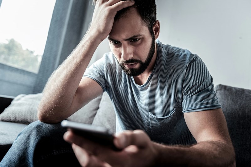 Man sitting on a couch looking stressed while holding a smartphone and resting his hand on his head.