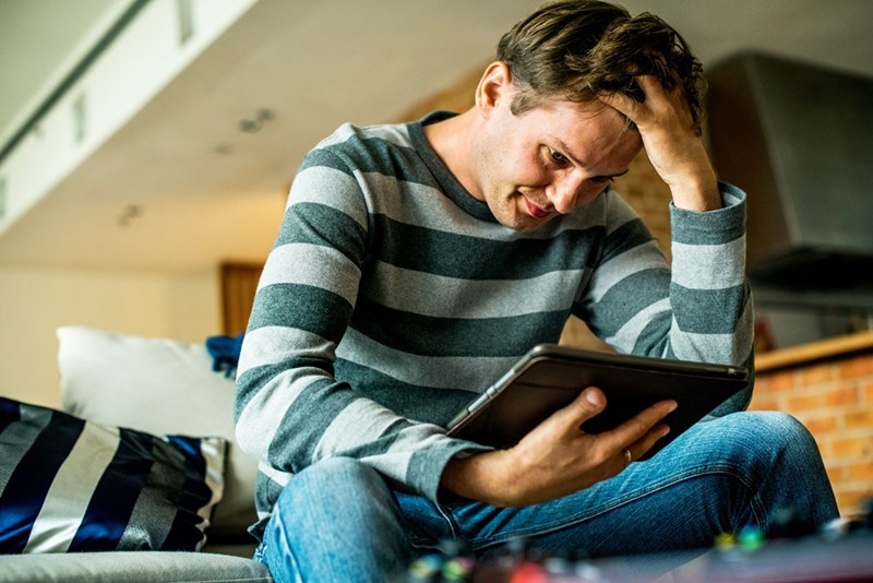 Man sitting on a couch looking frustrated while holding a tablet and resting his hand on his head.