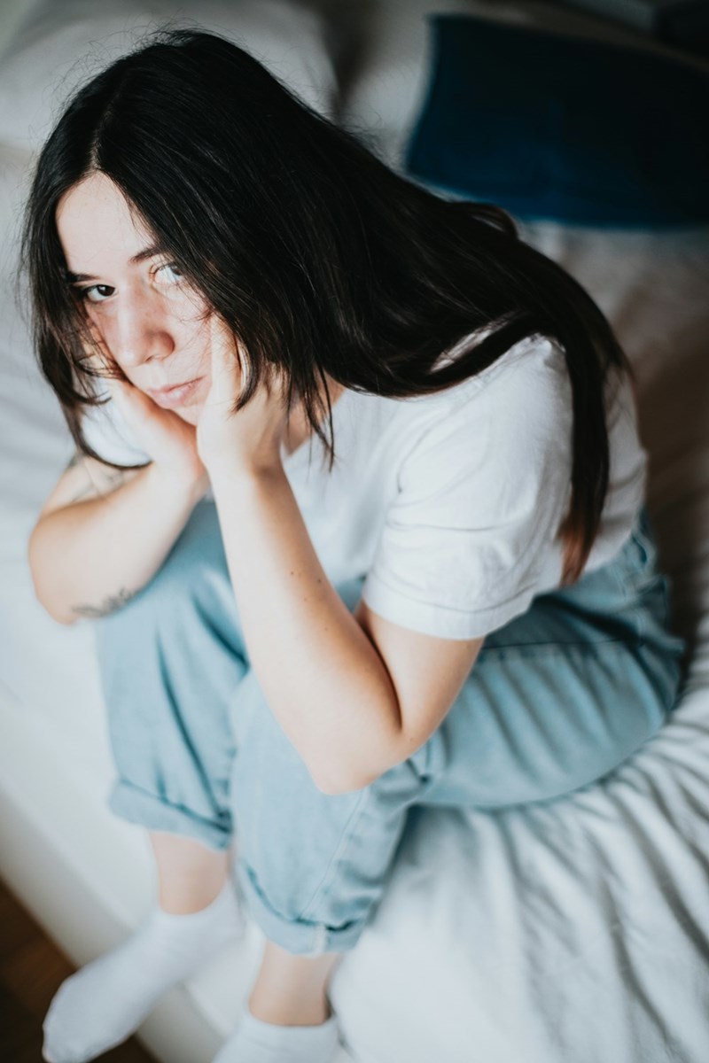 Young woman sitting on a bed with her hands on her face, looking tired and emotional.
