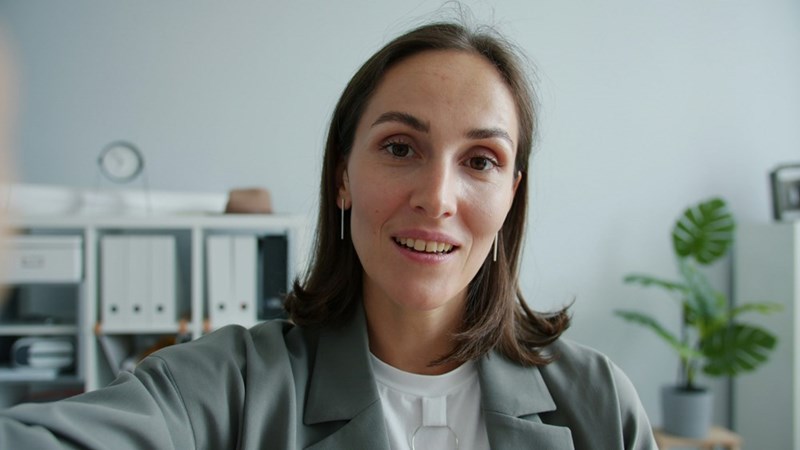 Professional brunette woman speaking to camera in a bright modern office with shelves and plants behind her.