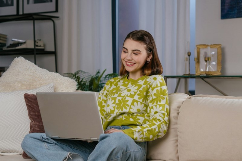 A woman smiles at her laptop while sitting cross-legged on her couch and wearing a cool, funky green floral sweatshirt