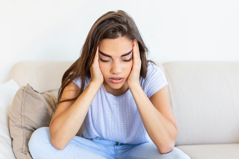 Woman with long brown hair sitting in a grey couch looking frustrated with her hands on her head.