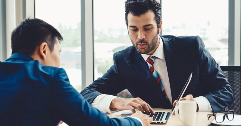 A manager in a business suit shows a member of his team something on his laptop in an office.