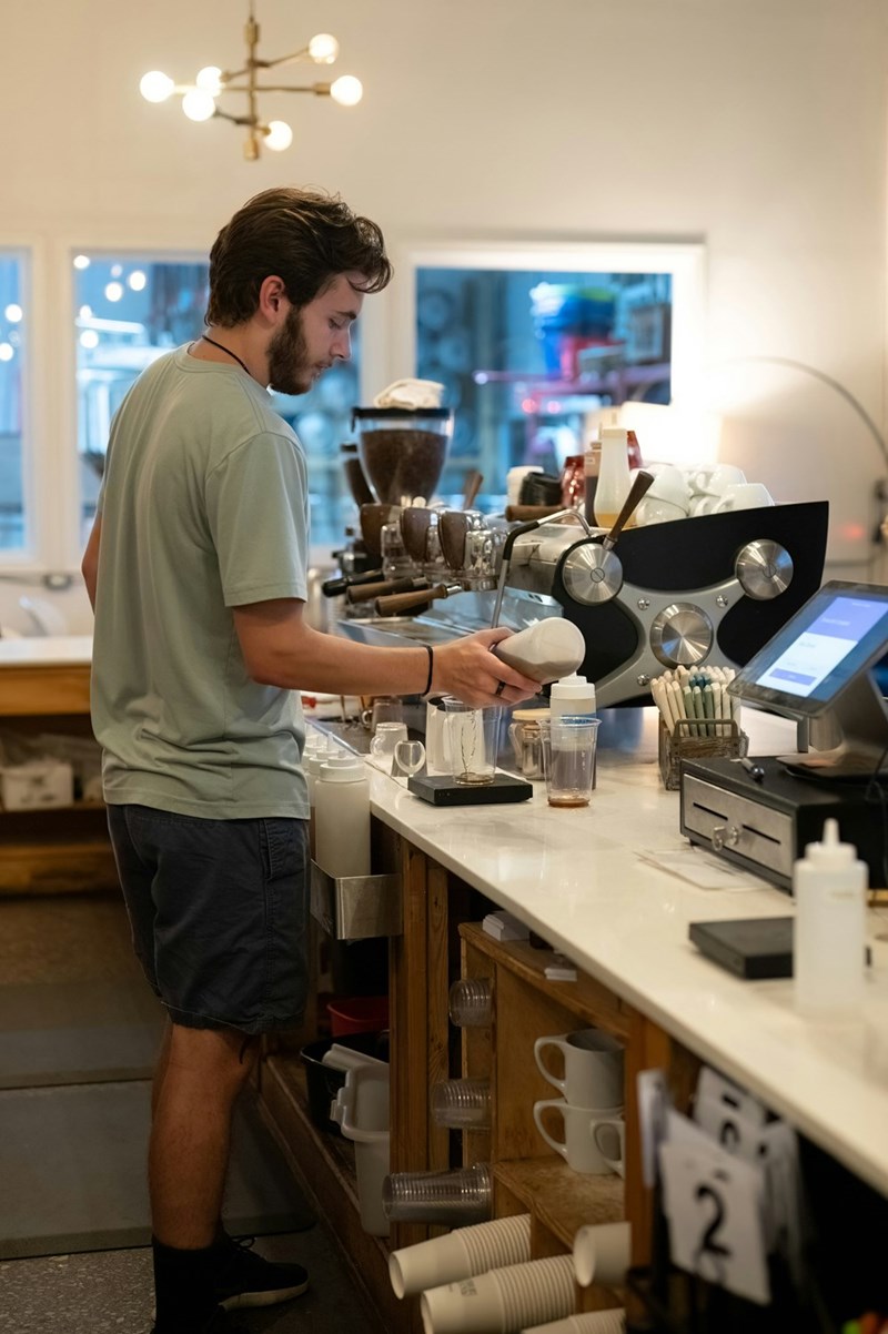 A young male barista stands in the kitchen preparing food for a customer.