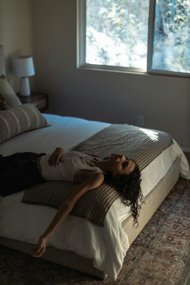 Conflicted woman dressed casually laying on her bed facing the ceiling with a window by her bed.