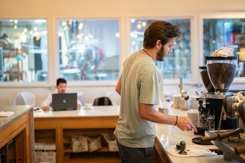 A young male barista takes orders from customers behind the counter as another customer sits with a laptop open in the background.