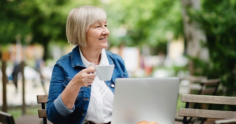 An older woman smiles with her coffee while sitting in front of a laptop at a small table outside a coffee shop.