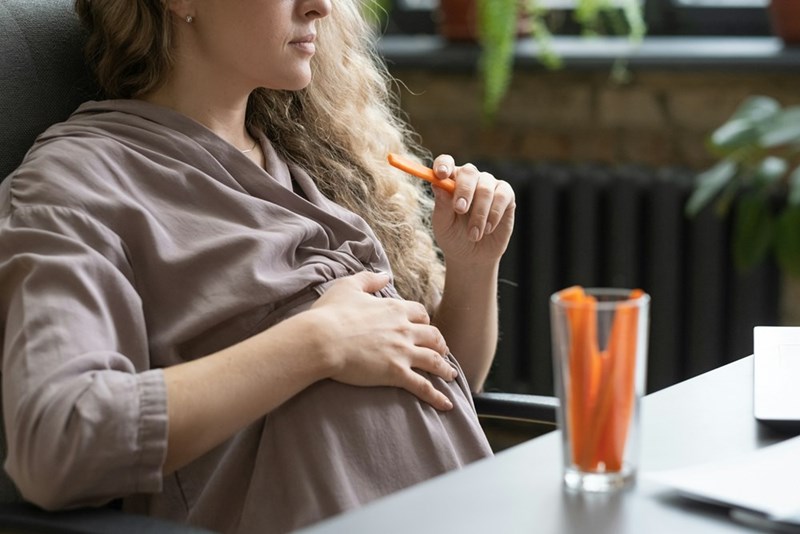 A pregnant woman eats a carrot as she touches her belly