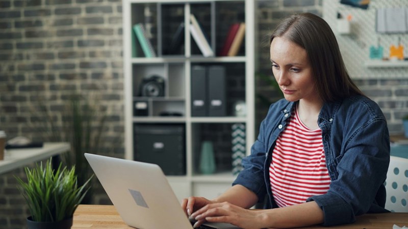 Woman working on a laptop at a wooden desk in a modern home office with shelves and plants in the background.