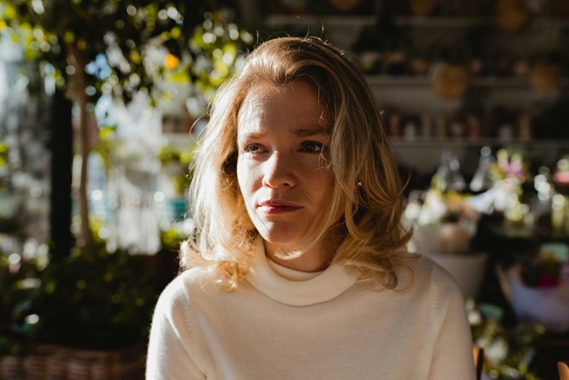 Blonde woman in a white turtleneck sitting indoors in warm sunlight with plants and shelves behind her.