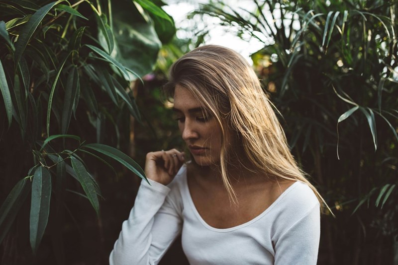 Blonde woman in a white long-sleeve top standing among tropical green plants in soft natural light.