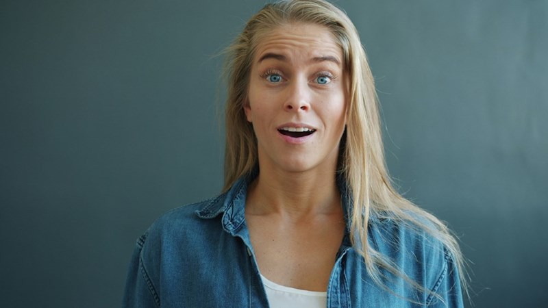 Blonde woman in a denim shirt with a surprised expression standing against a soft blue background.