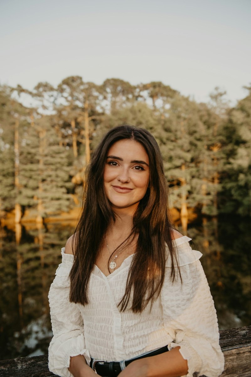 Brunette woman in a white off-shoulder blouse posing outdoors by a lake during golden hour.