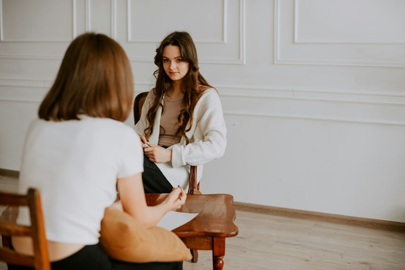 Two women sit in white looking at each other in conversation