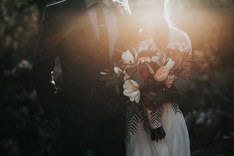 A bride and groom pose with the sun in the background and flowers in hand