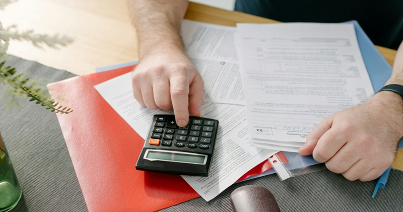 A man's hands type on a calculator on top of a pile of bills on a table