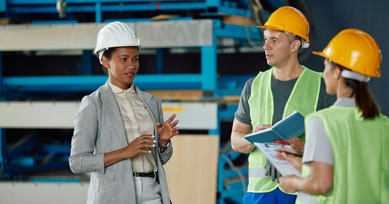 A warehouse supervisor speaks to employees, as they all wear hard hats