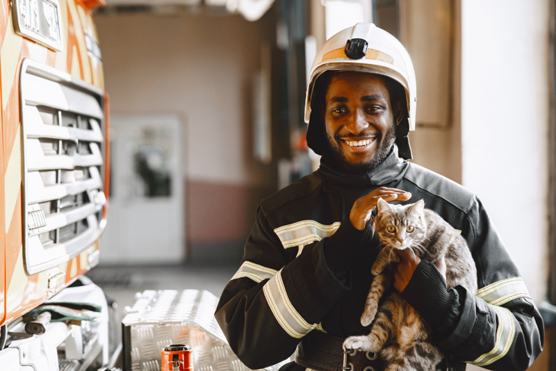 A firefighter holding a cat in his hands, smiling towards someone outside of frame.
