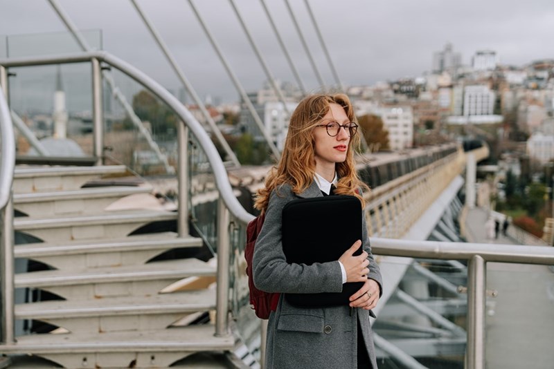 Woman with glasses holding a laptop case on a modern pedestrian bridge overlooking a city skyline.