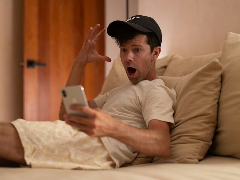 Man reacting in surprise to something on his smartphone while wearing a black baseball cap.