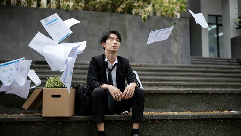 A young intern sits on the steps outside the office with his belongings after being fired, his papers fly thrown through the air around him.
