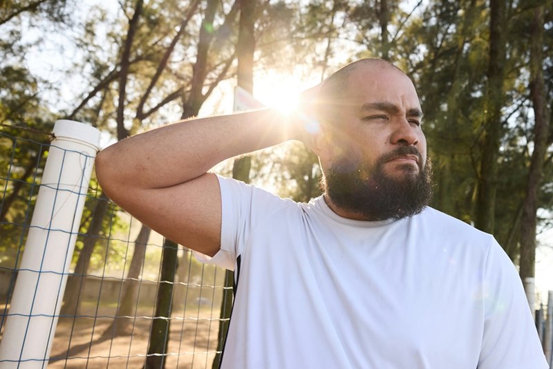 Bearded man outdoors in a white athletic shirt rubbing his neck in bright morning sunlight near trees.