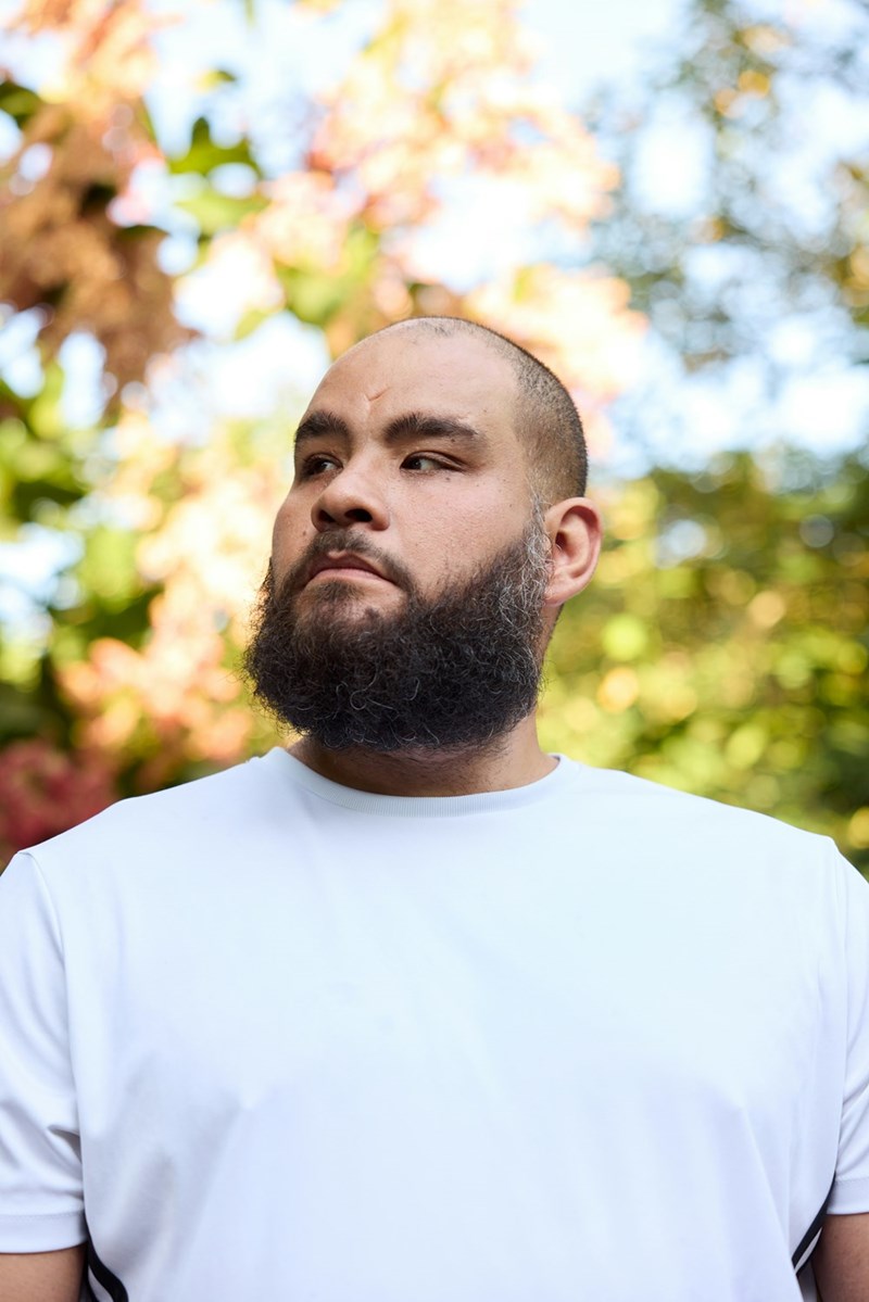 Bearded man in a white athletic shirt standing outdoors with autumn trees blurred in the background.