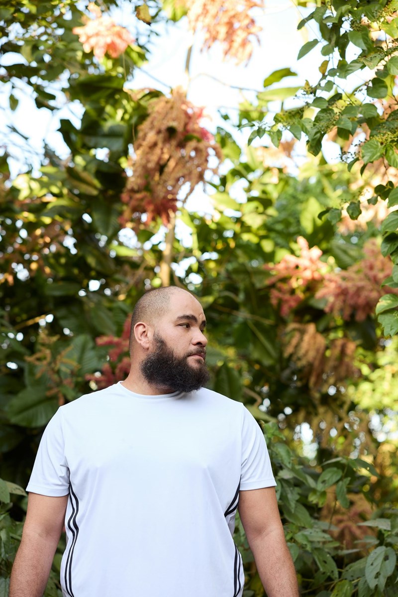 Bearded man in a white athletic shirt standing outdoors surrounded by dense tropical plants and trees.