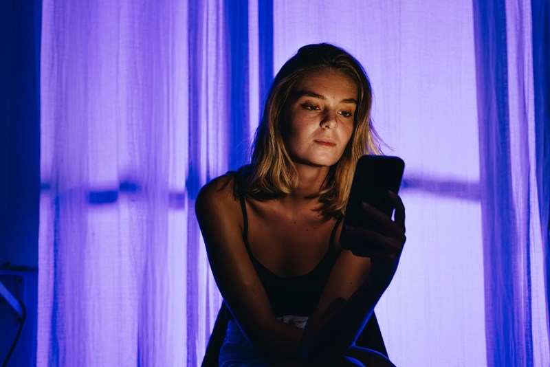 Woman using a smartphone at night indoors with blue ambient lighting and glowing curtains behind her.