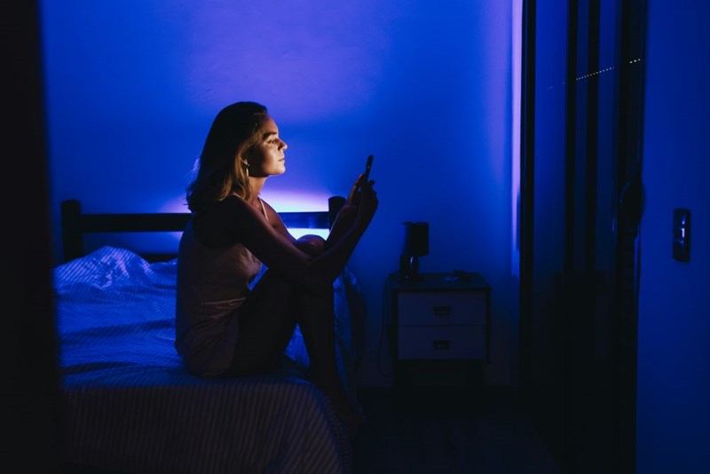 Woman sitting on a bed at night looking at a smartphone in a dark room lit by blue ambient light.