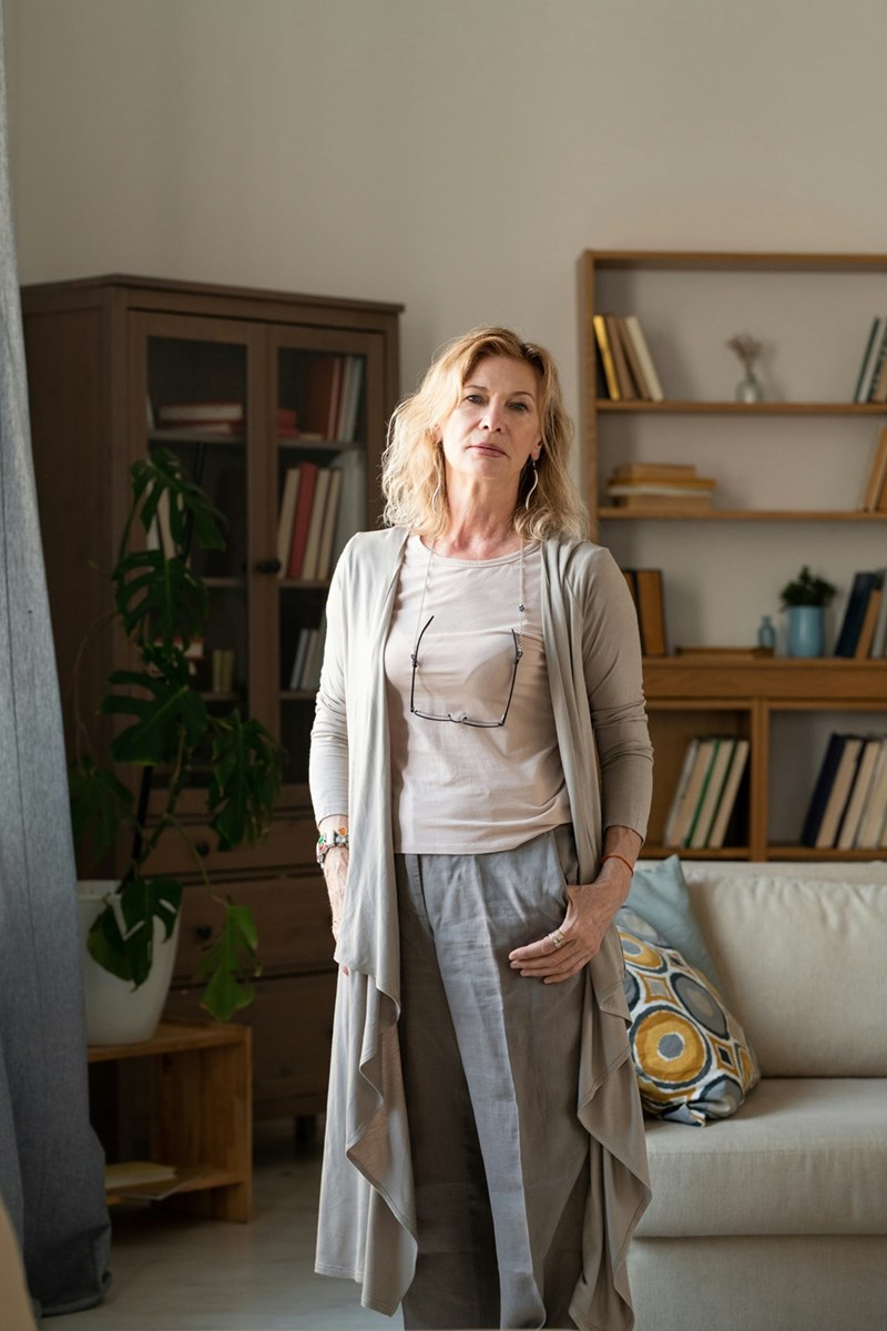 Middle-aged woman standing in a modern living room with bookshelves and neutral-colored clothing.