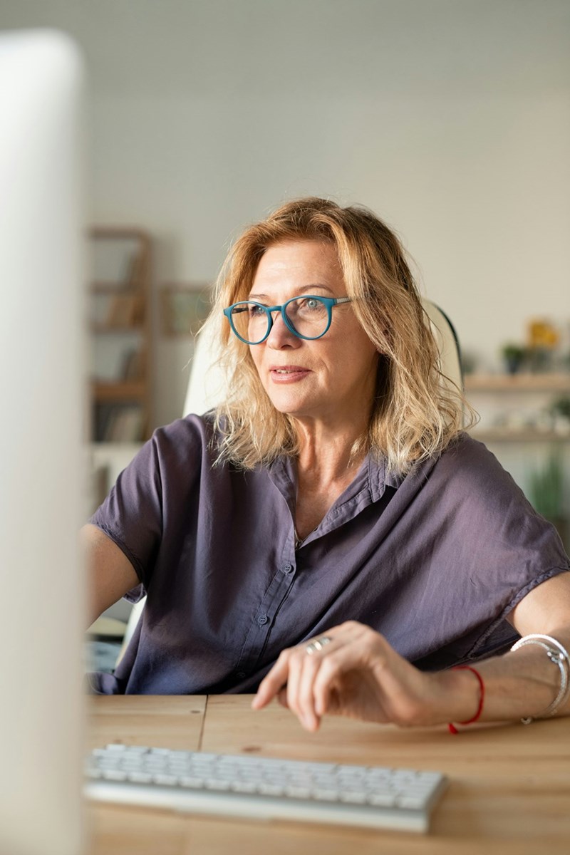 Middle-aged woman wearing blue glasses working at a desktop computer in a home office.